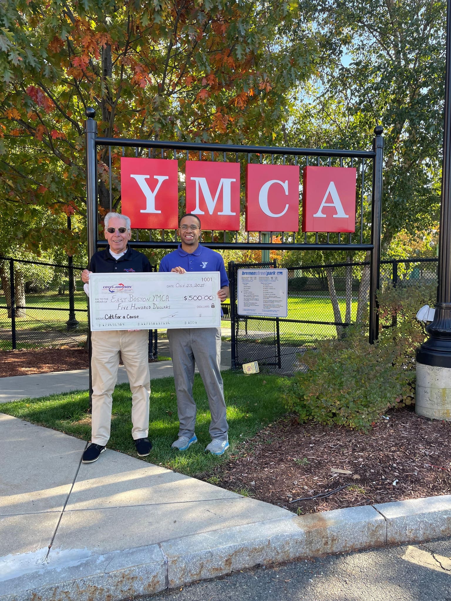 Cotti-Johnson HVAC representatives presenting a $500 check for the East Boston YMCA in front of the YMCA sign, showcasing community support through the 'Cotti For A Cause' initiative.